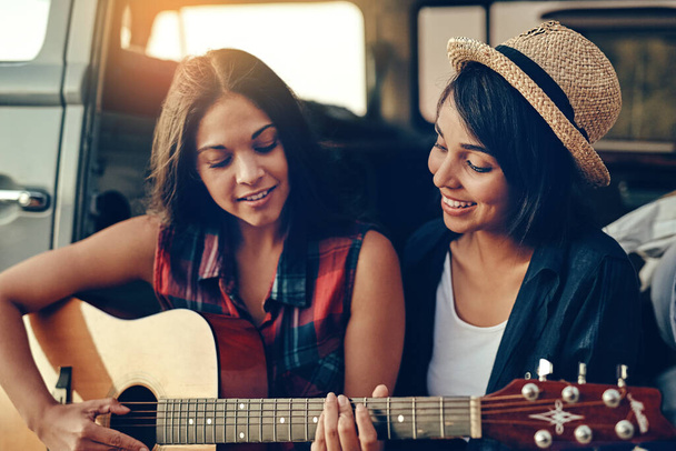 Hudba vytváří vzpomínky. Shot of a young woman playing a guitar on a road trip with her friend. - Fotografie, Obrázek