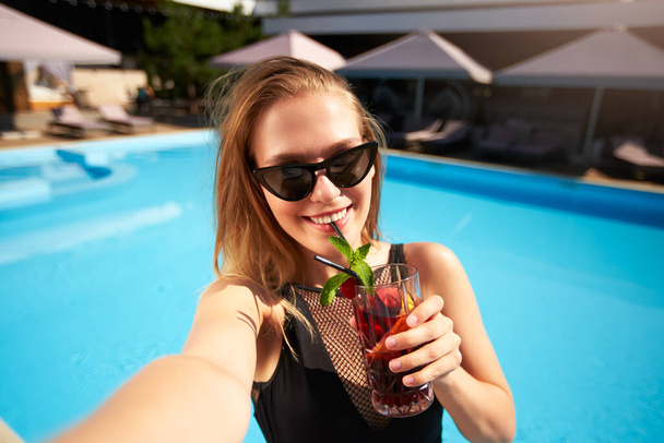 Travel blogger woman in bikini taking selfie photo with cocktail near swimming pool. Pov of lifestyle vlogger selfie from vacation on luxury resort. Female in swimwear drinks beverage on tropical spa. - Foto, Imagen