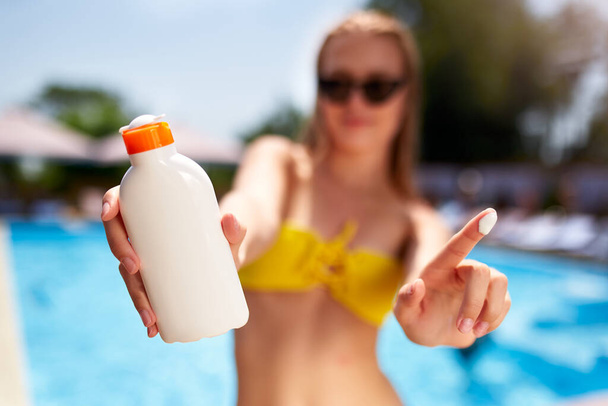 Closeup of smiling caucasian woman showing sunscreen oil bottle and using sun protection cream smear on finger. Pretty tanned girl wearing bikini swimsuit, straw hat and sunglasses on beach near sea. - Fotoğraf, Görsel