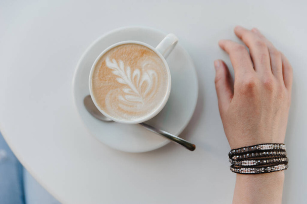 Top view. Morning mood. Cup of coffee with a pattern and a female hand with beads on a white table. High quality photo - Fotoğraf, Görsel