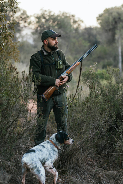 Hunter man with a gun standing outdoors with his dog while searching for a prey. - Photo, Image