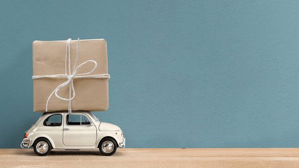 Retro white toy car delivering gift box wrapped in kraft paper atop on blue background. Copy space - Photo, Image