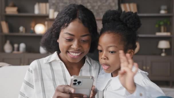 Cute little african kid child daughter having fun with multiracial mum at home using smart phone typing swipe screen play mobile game app on modern gadget cellphone looking at telephone sit on sofa - Footage, Video