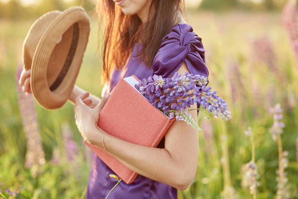 Kobieta w letnim polu z bukietem dzikich kwiatów - łubin, kapelusz i różowy notatnik. Bliskość natury, koncepcja samopoznania. Relaks i dobre samopoczucie koncepcja. Słoneczny dzień, dobry nastrój. - Zdjęcie, obraz