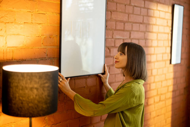 Woman hangs a photo frame on brick wall at home - Photo, Image