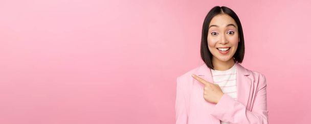 Портрет азиатской деловой женщины, продавщицы в костюме указывая пальцем влево, показывая баннерную рекламу, улыбаясь и глядя профессионально, розовый фон - Фото, изображение