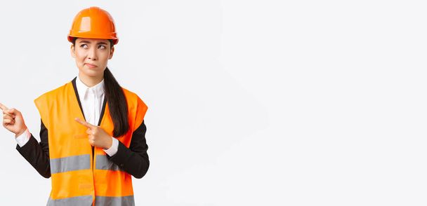 Building, construction and industrial concept. Indecisive asian female architect being unsure, wearing reflective clothing and helmer, smirk as pointing and looking upper left corner puzzled - Photo, Image