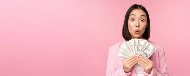 Finance, microcredit and people concept. Happy smiling asian businesswoman showing dollars money, standing in suit against pink background - Fotó, kép