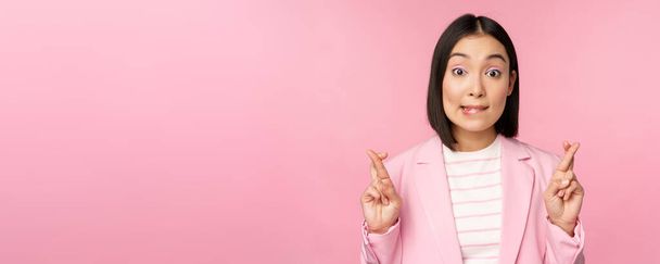 Счастливая удачливая деловая женщина, азиатская корпоративная леди желает, загадывает желание, надеется на смесь и молится, стоя в костюме на розовом фоне - Фото, изображение