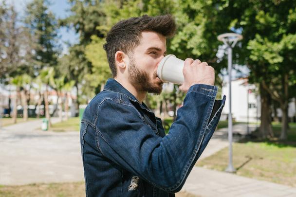 Красавчик в синей джинсовой куртке отводит взгляд, когда пьет кофе. Человек думает о своей жизни в летний день. - Фото, изображение