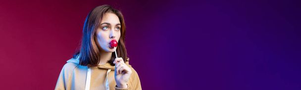 Egy kapucnis nő portréja, a lányok egy piros kerek nyalókát nyalogatnak gyönyörű sminkkel, lila trendi háttérrel a stúdióban.. - Fotó, kép