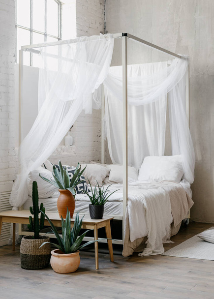 Vertical shot of white baldachin over comfortable bed in boho chic bedroom. Morning light in hygge room with potted house plants on wooden bench. Concept of apartment with scandinavian interior style - 写真・画像