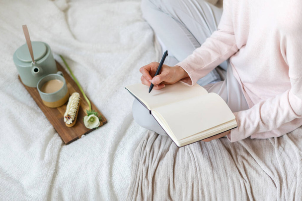 Cropped shot of woman in pyjamas makes notes in diary journal with blank pages. Female sits on bed with breakfast tray, enjoy morning at home. Blogger create publication in notebook, high angle view - Photo, image