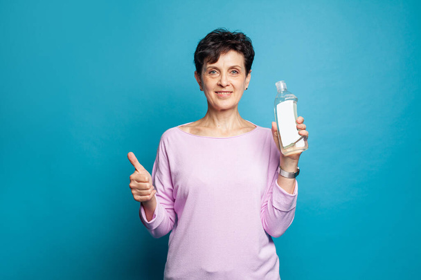 Щаслива дружня зріла жінка тримає пляшку для прибирання і показує великий палець на синьому фоні - Фото, зображення