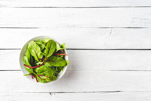 Overhead shot of fresh green salad with spinach, arugula and beetroot leaves in bowl on white wooden background with copyspace - 写真・画像