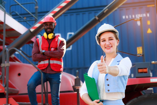 Portrait of a beautiful Caucasian female manager in a white hard hat, Container Loading Supervisor. Behind it is a blurred image of an African American man. Port concept of import and export goods. - Фото, изображение