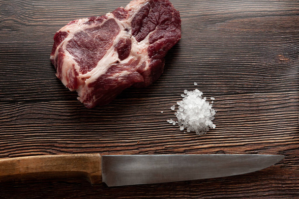 een rauw stuk varkensvlees op een donkere houten achtergrond ernaast ligt een mes en zout - Foto, afbeelding