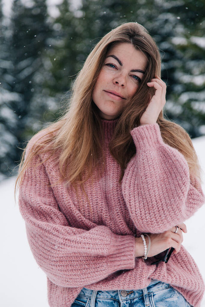 A young, beautiful, long-haired girl in a pink sweater poses in a winter forest in the mountains - Fotoğraf, Görsel