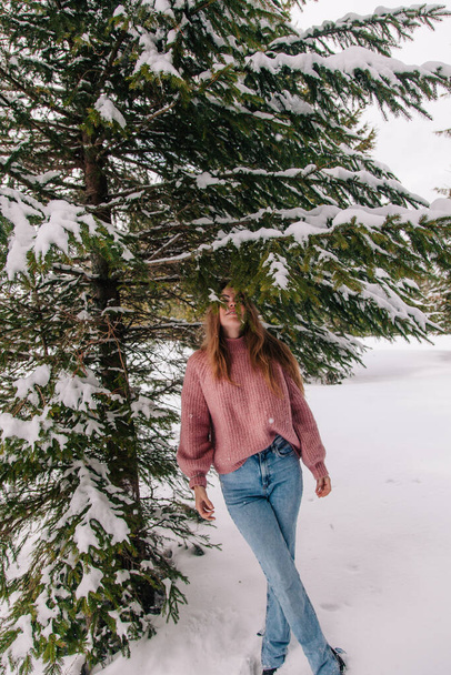 A young, cheerful girl sprinkled with snow under the branches of a spruce tree in the mountains - Photo, image