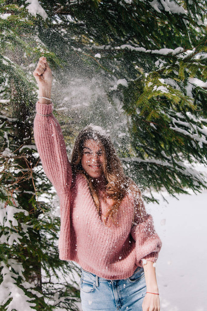 A young, cheerful girl sprinkled with snow under the branches of a spruce tree in the mountains - Foto, imagen