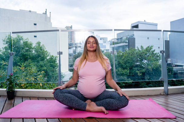 front view young caucasian woman with closed eyes pregnant on the balcony at home doing yoga and meditation on a pink mat, healthy life and motherhood concept. - Фото, изображение