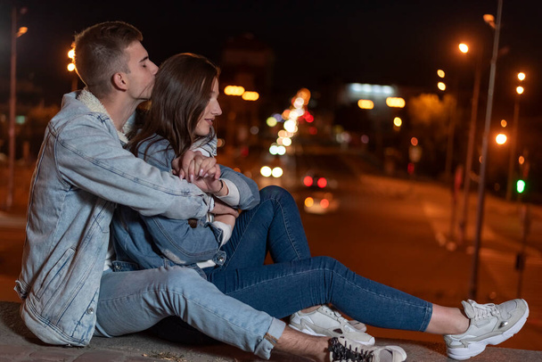 Jeune couple caucasien est assis sur le trottoir s'embrassant dans le contexte des lumières de la ville de nuit. Date et relation. - Photo, image