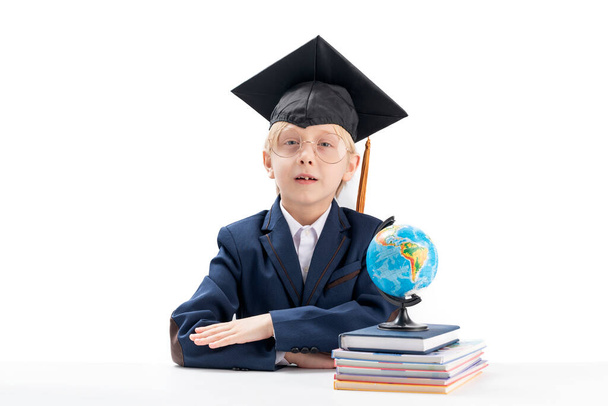 Niño de escuela primaria con gafas grandes y sombrero de estudiante se sienta a la mesa, cuadernos y glob. Aprendizaje rápido. Aislado sobre fondo blanco - Foto, Imagen