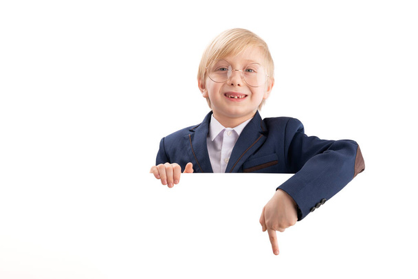 Boy in school uniform and large glasses with blank white poster board peeking. Schoolboy points his finger at something. Copy space - Фото, изображение