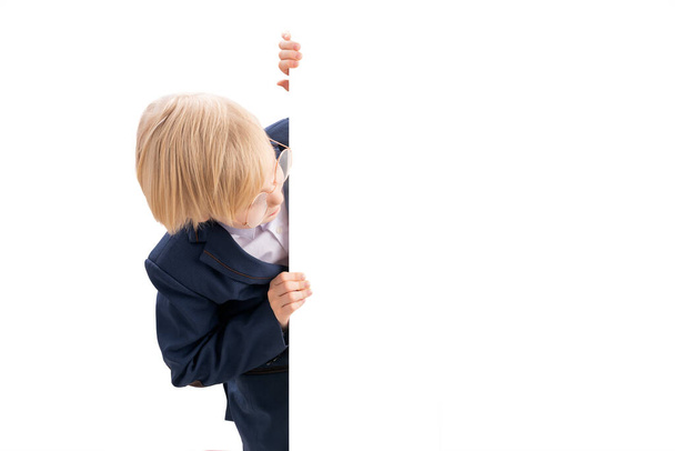 Koulupoika kurkistaa tyhjästä tyhjästä. Vaalea poika koulupuvussa tyhjällä valkoisella julisteella. Kopioi tila - Valokuva, kuva