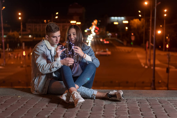愛を持っている学生のカップルは、彼らの手の中にガーランドと夜の街の背景に座っている。夜市灯. - 写真・画像