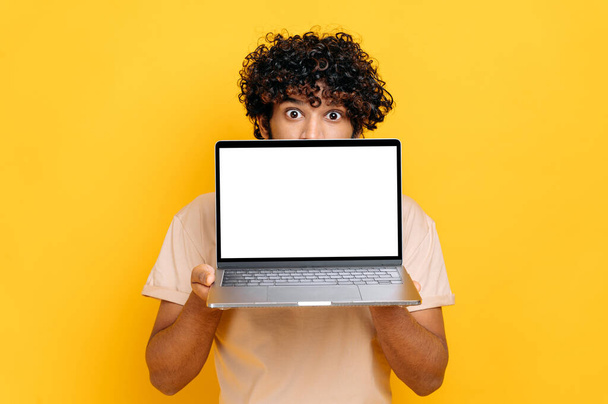 Удивленный возбужденный индиец или араб, выглядывающий из-за ноутбука, смотрит удивлённый камерой, стоит на изолированном оранжевом фоне, держит открытый ноутбук с чистым белым макетом экрана, копирует пространство - Фото, изображение