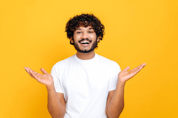 Joyful positive indian or arabian guy with curly hair, wearing white t-shirt, looks at camera happily, spreading his arms to the sides, standing over isolated orange background, looks at camera, smile - Fotó, kép