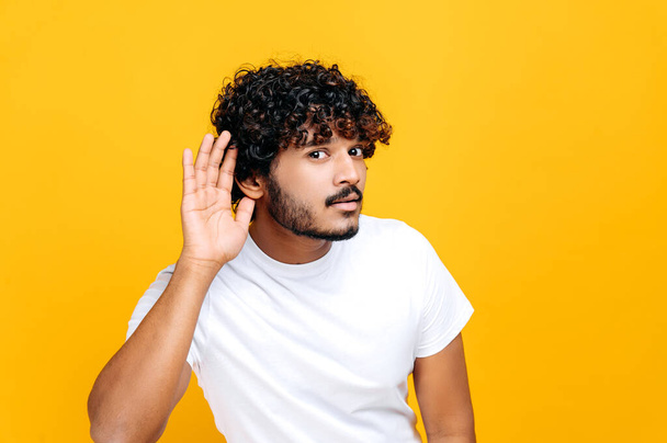 Positive Indian man, wearing basic t-shirt, holds hand near ear concentrated listening rumor, hearing gossip, standing on isolated orange background. Deafness, gossip concept - Photo, image