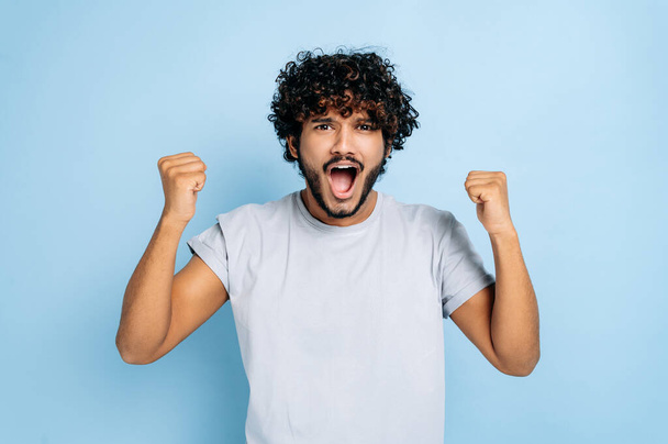 Win, triumph. Excited amazed indian or arabian guy, in t-shirt, rejoices in success, victory, win, gesturing with fists, looking at camera, smiling, standing on an isolated blue background, shouting - Foto, Imagem