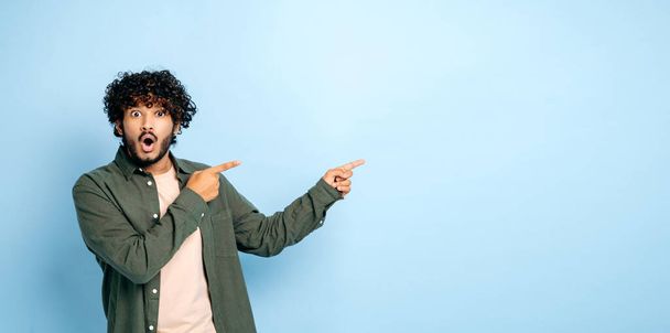 Panoramic photo of amazed shocked indian or arabian guy in casual wear, points fingers to the side at mock-up empty space, stands on isolated blue background, looks surprised at the camera, astonished - Valokuva, kuva