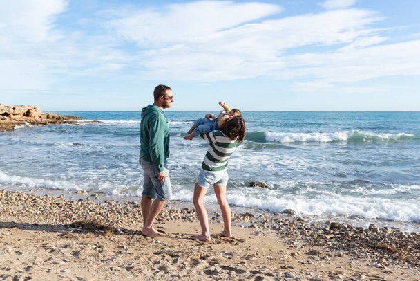 Matka a otec si hrají se svou dcerou na pláži. Matka drží své batole, zatímco ji líbá a lechtá, a otec se na ně dívá. Láska a smích na pláži.  - Fotografie, Obrázek
