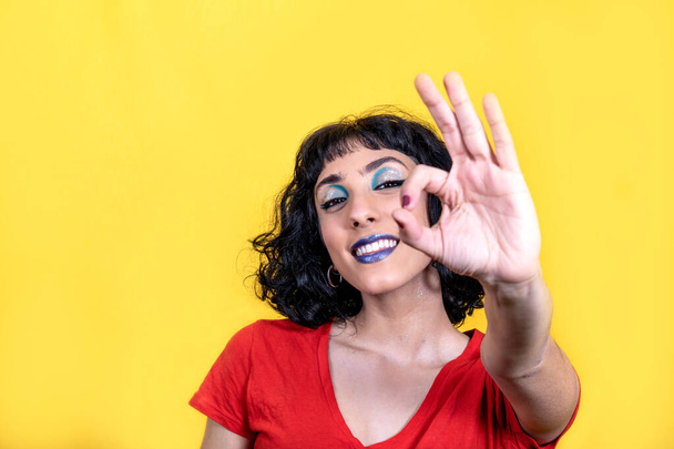 Smiling young woman in red t-shirt is showing ok sign with fingers. Woman portrait with trendy look and bright colors on yellow background. - 写真・画像