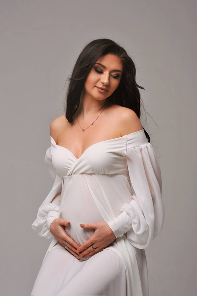 Portret pięknej ciężarnej kobiety w białej sukience siedzącej na krześle i trzymającej jej brzuch w ciąży, odizolowanej na białym tle. Koncepcja szczęśliwego macierzyństwa i rodzicielstwa - Zdjęcie, obraz