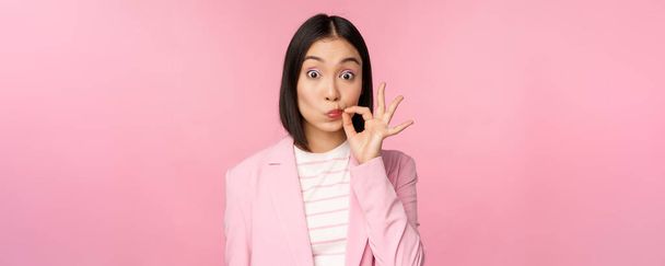 Portrait of asian corporate woman showing mouth seal, close shut lips on key gesture, promise keep secret, standing over pink background in suit - Photo, Image