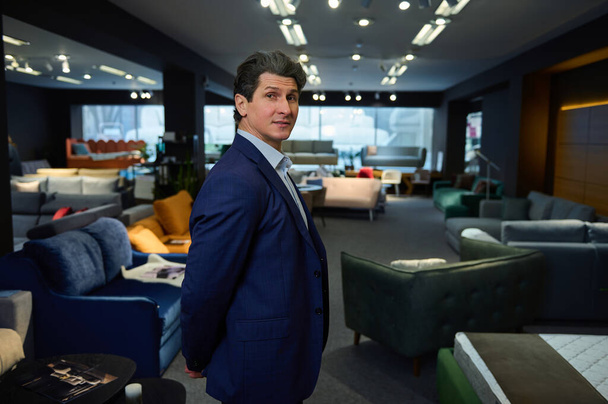 Confident business owner, entrepreneur, handsome imposing Caucasian mature man in the showroom of a furniture store with a wide range of sofas, couches and upholstered furniture - Foto, afbeelding