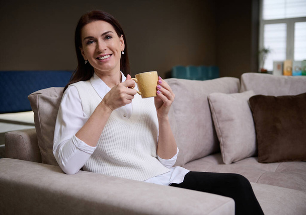 Bella donna europea serena seduta sul divano e rilassata, che si gode una tazza di caffè mentre sceglie un comodo divano nello showroom del negozio di mobili. Home interior design e concetto di miglioramento - Foto, immagini
