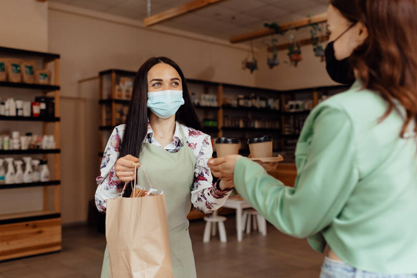 Ένα νεαρό κορίτσι με μάσκα αγοράζει καφέ για να πάει. Ιδιοκτήτης μικρής επιχείρησης που φοράει ιατρική μάσκα στον πάγκο σε καφέ ή μικρό κατάστημα. έννοια της υγειονομικής περίθαλψης και της τεχνολογίας κατά τη διάρκεια της πανδημίας coronavirus - Φωτογραφία, εικόνα