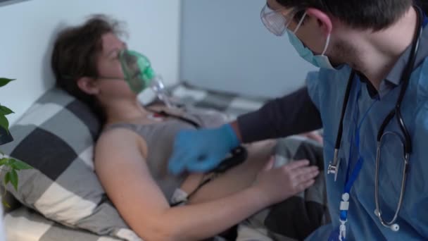 Mavi üniformalı bir erkek doktor ve oksijen maskeli bir kadın hasta evde yatıyorlar. Doktor, koronavirüs sırasında karantinaya alınmış, solunum ve kalp rahatsızlığı olan kadınlara tıbbi muayene yapıyor. - Video, Çekim