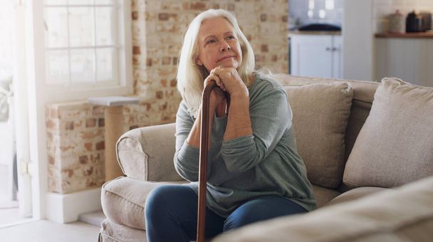 Me pregunto qué podría hacer hoy. Foto de una mujer mayor sentada sola en el sofá en casa y mirando contemplativa mientras sostiene su bastón. - Foto, imagen