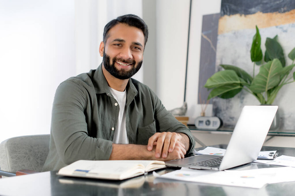 Pozytywny pewny siebie Indianin lub arabczyk korzystający z laptopa w nowoczesnym biurze. Portret odnoszący sukcesy biznesmen, freelancer, manager, siedzi w miejscu pracy przed laptopem, patrząc w kamerę, uśmiechnięty przyjazny - Zdjęcie, obraz