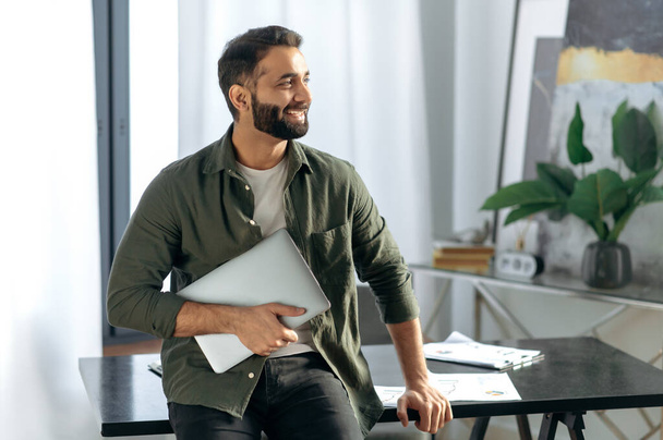 Portrait of an attractive modern Indian or Arabian successful entrepreneur or freelancer in casual clothes, stands in his modern office near the table, holds a laptop, looks away, smiling - 写真・画像