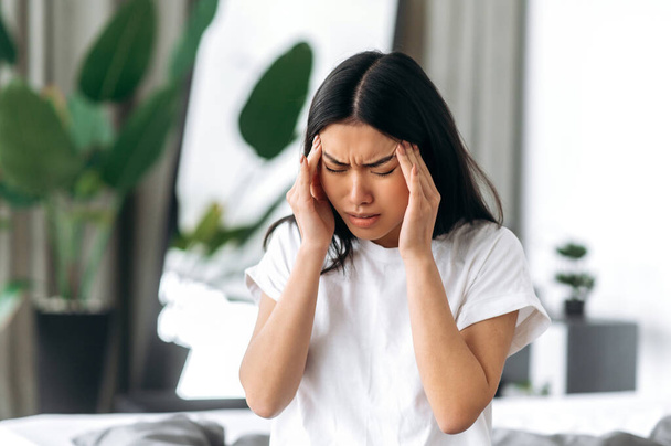 Upset Asian exhausted girl in white t-shirt, sits in a room on a bed, massages her temples, closed her eyes, suffers from headache, migraine, overwork, head spasm, needs treatment, rest and sleep - Фото, изображение