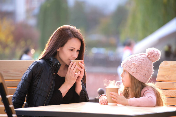 Η νεαρή μητέρα και η κόρη της περνάνε καλά μαζί στο καφέ του δρόμου με ζεστά ροφήματα την ηλιόλουστη φθινοπωρινή μέρα. Η ευτυχία στις οικογενειακές σχέσεις - Φωτογραφία, εικόνα