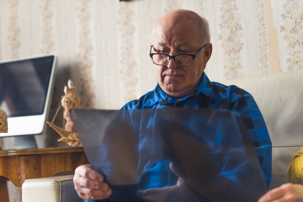 Zaniepokojony stary pomarszczony łysy mężczyzna w niebieskiej flanelowej koszuli i czarnych okularach patrzący na jego prześwietlenie i martwiący się o jego zdrowie. - Zdjęcie, obraz