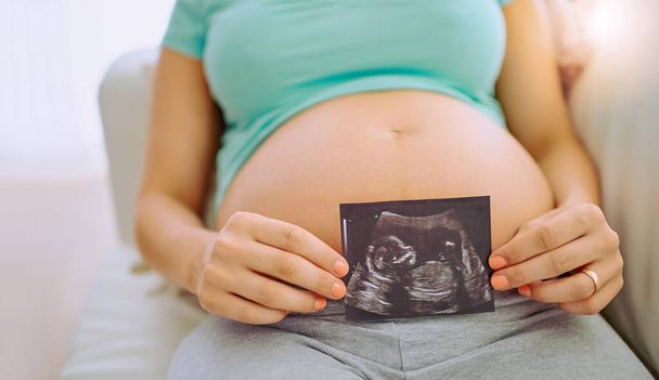 Ontmoet het kleine mensje in mijn buik. close-up shot van een zwangere vrouw met een echo foto tegen haar buik thuis. - Foto, afbeelding